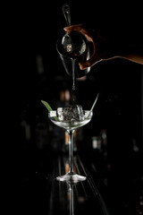 Female bartender's hand holds mixing cup with filter and pours thin stream of clear liquid into martini glass at bar counter