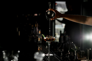 Selective focus of backlited martini glass in which female bartender pours liquid from steel shaker cup