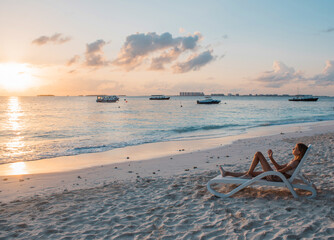 A girl lies on a deck chair on the beach by the ocean on a tropical island and looks at the sunrise and the beautiful sky in the rays of the rising sun.  Maldive Islands. Travel, vacation content