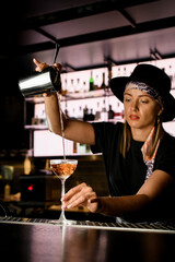 Female bartender carefully pours cocktail from shaker with strainer to drinking glass at bar counter