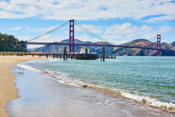 Waves crashing against beach with pier and small shack beneath Golden Gate Bridge