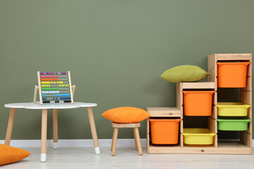 Table with chair, shelves and abacus toy near olive wall in playroom. Stylish kindergarten interior