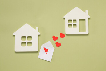 Long-distance relationship concept. Envelope with paper hearts between two white house models on green background, flat lay