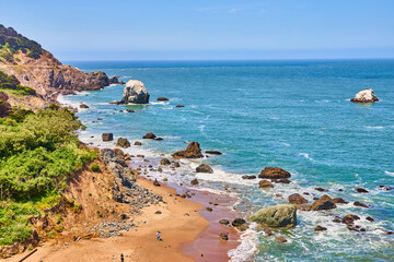 Fototapeta na wymiar Sunny beach near rocky coastline with people near waves on gorgeous blue sky day