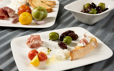Multiple appetizers on white plates with roasted tomatoes, olives, prosciutto, olive oil, bread