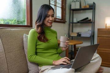 Young successful professional Asian businesswoman in casual clothes sits on a sofa, reads a book, writes notes in a notebook while working on a laptop computer at home office in the living room.