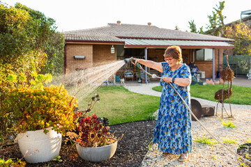 Older female in her backyard watering garden with hose in evening light