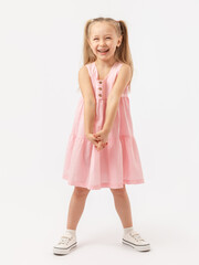 A cheerful girl with long hair in a pink dress and sneakers stands on a white background and laughs sincerely.