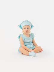 A cute 2-year-old toddler girl in a blue dress and a kerchief sits half-sideways on the floor and looks attentively to the side on a white background.
