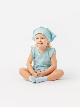 A cute 2-year-old toddler girl in a blue dress and a scarf is sitting on the floor smiling and looking up attentively on a white background.