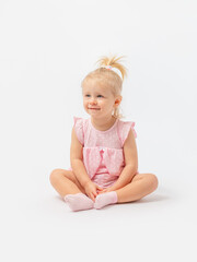 A cute blonde girl 1-2 years old in a pink dress is sitting on the floor smiling and looking away on a white background. Copy space.