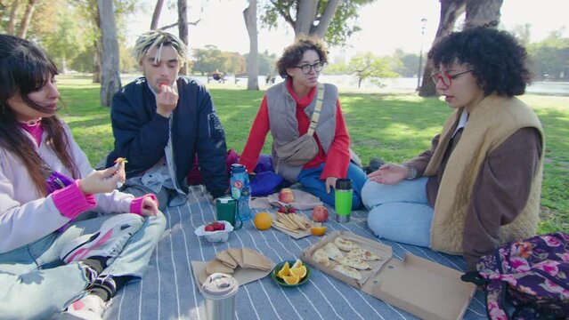Young cheerful friends enjoying fresh fruit and snacks, talking and having fun on a picnic in the park after study day