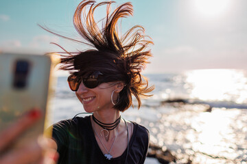 woman with hair blowing in the wind taking a selfie by the sea. Beautiful girl smiling while looking at camera outside - fashionable lifestyle and beauty concept