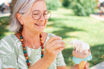 Smiling senior woman sitting outdoors in the park eating an ice cream cone. Elderly caucasian...