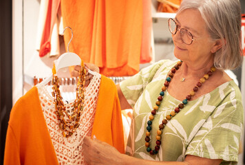 Carefree senior woman looking for sale in a store selecting colorful clothes in orange color enjoying shopping, consumerism sales customer retail concept