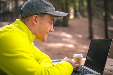 Beautiful caucasian boy influencer working on laptop sitting outdoors in the forest. Hipster young man traveler working distantly while enjoying nature landscape during vacations - obrazy, fototapety, plakaty