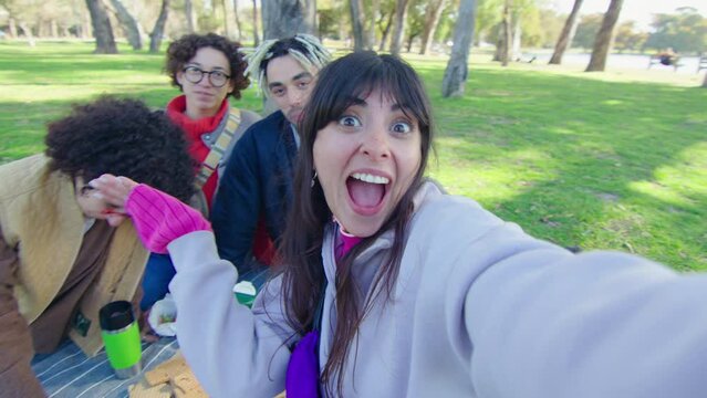 Young excited friends waving, making fun, eating snacks and posing together for camera while filming social media video or taking selfie on picnic in park. POV shot