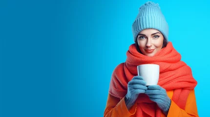 Foto op Plexiglas Beautiful young female in a hat, scarf and gloves with cup of hot cocoa on color background. Woman in colorful winter clothes holding a cup of hot drink standing on the blue background with copy space © pijav4uk