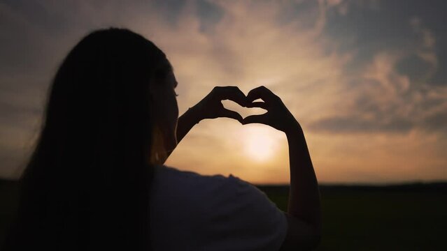 Happy girl figure from fingers. Heart with hand symbol of love and freedom. Man dream. Girl outdoors in park silhouette at sunset. Heart fingers hand figure in nature. Happy family and freedom concept