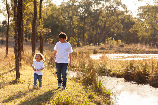 Aboriginal brother and sister walking together by creek in australian paddock