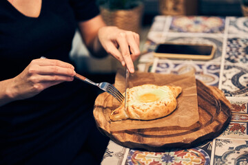 Women's hands cut khachapuri in Ajarian style with an egg on a wooden stand. Traditional Georgian pastries - khachapuri in Adjarian style with cheese and raw yolk. High quality photo
