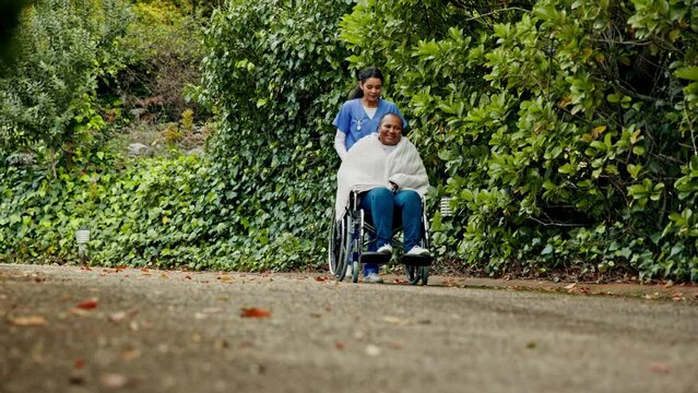 Woman, nurse and patient walking with wheelchair in nature for outdoor healthcare, support or trust. Female doctor or medical caregiver pushing person with a disability in elderly care or nursing