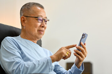 Pleased senior man browsing mobile apps, chatting with relatives online on mobile phone