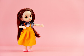 child toy doll with dark hair in an orange dress standing on a pink background. Plastic children's...