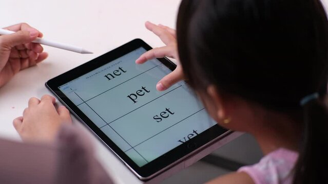 Happy little Asian daughter talking with mother using digital tablet to do homework or study online. Happy young girl and adult sitting at table learning on iPad together. Back to school.