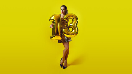A slender young tanned woman in a shiny dress holds the number 13 folded from golden inflatable balloons. Holiday, birthday. Number 13. Model posing on a golden background.
