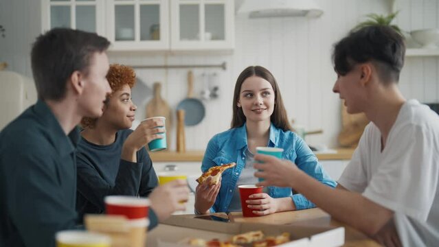 Company young friend having lunch eating pizza sitting on kitchen table talking smiling at home. Pastime, leisure together, friendship, lifelong friends concept. Multi ethnic people happy together.
