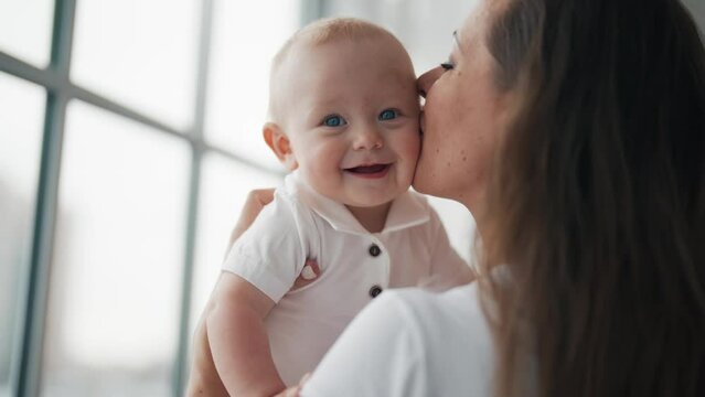 Portrait of smiling happy adorable baby standing all fours. Mom kissing little kid playing together at home. Babysitting, maternity leave motherhood babyhood parenting, mommy love and care concept.