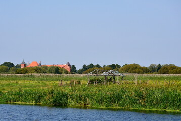 A view of a coast or a bank of a shallow yet vast river located next to a lush farmland, a ruin of a wooden hut or house and a massive medieval castle seen in the distance spotted on a sunny day