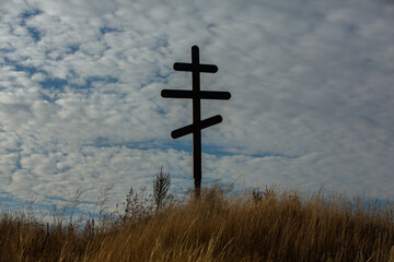 Cross on top of a hill against a blue sky with white clouds. Silhouette of a cross in the field at sunset with dramatic sky.