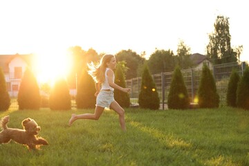 Beautiful girl running with cute Maltipoo dog on green lawn at sunset in backyard