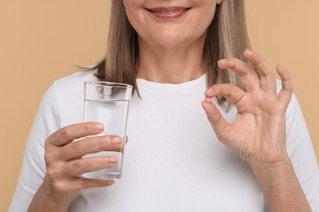 Senior woman with glass of water and pill on beige background, closeup