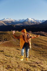 Full-length woman running up the hill to the camera and looking at the mountains in a yellow raincoat and jeans in the autumn happy sunset hiking trip mountains in the snow, freedom lifestyle 