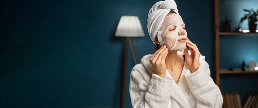 Beautiful Woman Applying In Sheet Face Mask Looking At Mirror. Procedure For Skin Moisturizing. Skincare And And Beauty Treatments Concept