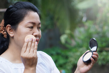 Asian middle aged woman is making up, using puff to apply powder on her face, looking at portable...