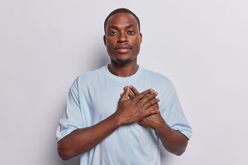 Waist up shot of dark skinned man heartedly touching chest dressed in casual blue t shirt expresses gratitude thanks God for something pleasant dressed in casual t shirt isolated over white background