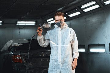Automotive service worker in protective gear expertly applying color paint in to car's bodywork with spray gun or respirator painting in chamber workshop. Car paint service for scratch refinish. Oxus