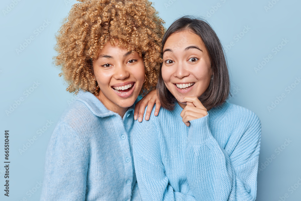 Wall mural Portrait of cheerful women smile broadly stand closely to each other dressed in casual jumpers look directly at camera isolated over blue background. People friendship and happiness concept. - Wall murals