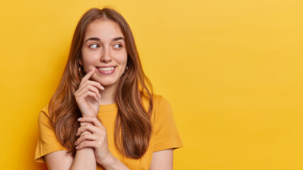 Horizontal shot of good looking young woman with long hair keeps hand near corner of lips concentrated aside smiles gently dressed in casual t shirt isolated over yellow background copy space for text