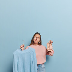 Upset long haired woman focused above with discontent expression holds sandals in one hand and jumper on hanger chooses what to wear for walk poses against blue background copy space overhead