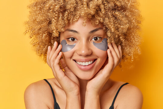 Beauty and cosmetology concept. Studio close up of young pretty cheerful smiling African american female standing on yellow background applying grey patches to reduce wrinkles with hands on cheeks