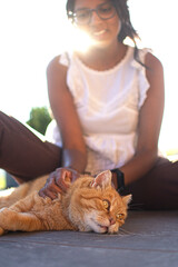 Smiling girl sitting caresses cat with orange fur lying down, green eyes watch carefully in front of him.