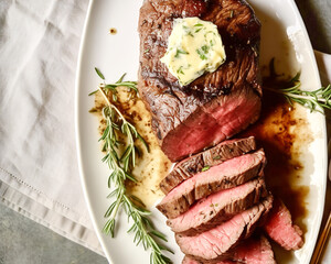 Delicious roasted beef tenderloin with herb butter on white plate garnished with fresh rosemary. Holiday meal for family dinner celebrations - obrazy, fototapety, plakaty