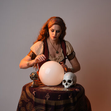 close up portrait of beautiful red haired woman wearing a medieval fantasy fortune teller costume, looking into crystal ball reading the future at seance table. isolated on studio background 