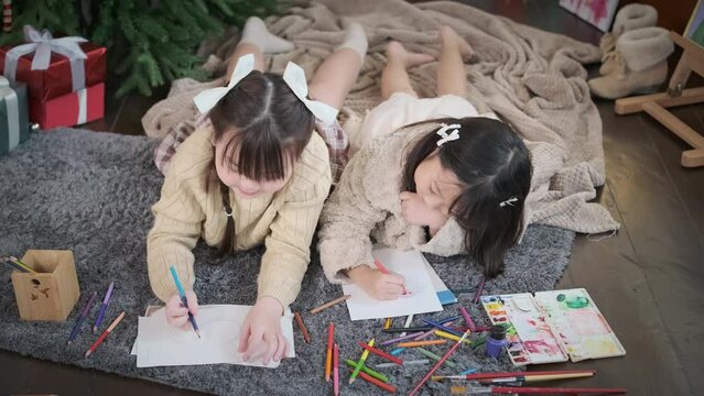 Cheerful little sisters making greeting card for New Year and Christmas while lying on floor in decorated living room