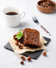 Pieces of chocolate brownies with hazelnuts, mint and salted caramel on baking paper on a light background with a cup of coffee.